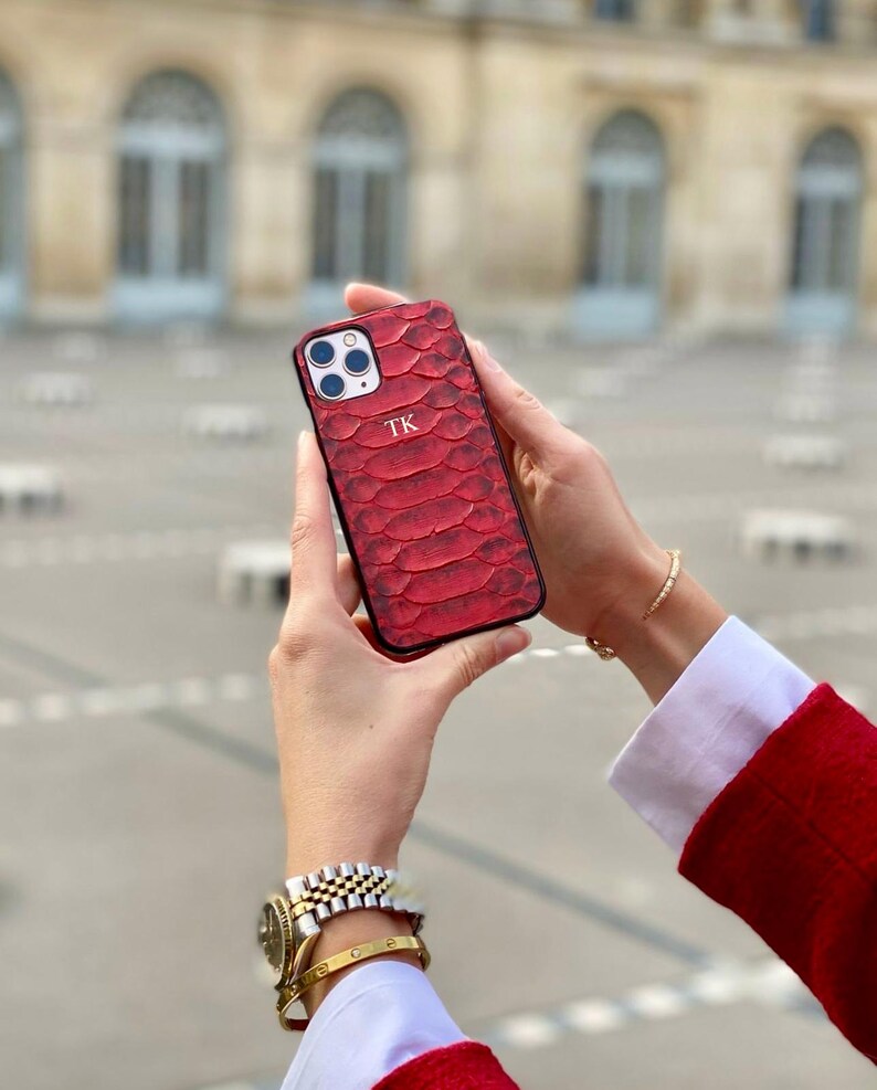 May include: Red textured phone case with the initials "TK" in white. The case has a snake-skin pattern and a black border. The phone case is held in front of a blurred background.