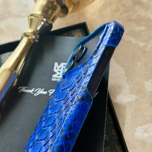 May include: A blue snakeskin-textured phone case. The case has a dark blue border and a textured surface. The case is resting on a black box with the text "Thank You".