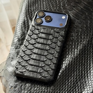 May include: A black phone case with a textured, scaly pattern, possibly leather or a similar material. The case is on a surface with a matching texture. The phone's camera lenses are visible.