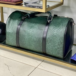 Puede incluir: Una bolsa de viaje de cuero verde con ribetes de cuero negro y un patrón de piel de serpiente. La bolsa tiene un asa superior y un cierre de cremallera.