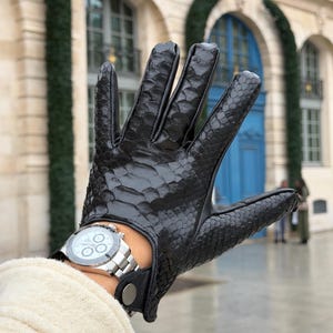 May include: Black leather glove with a snakeskin pattern. The glove is worn with a silver wristwatch. The glove has a button closure at the wrist. The background is a blurred outdoor scene.