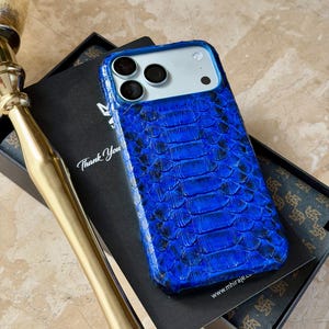 May include: A blue snakeskin-textured phone case with a silver camera module. The case is on top of a black box with the words "Thank You" and a website address. A gold-colored decorative object is in the foreground.