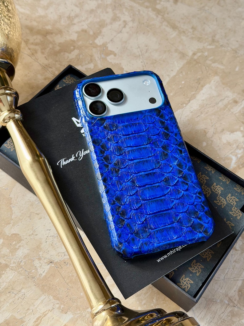 May include: A blue snakeskin-textured phone case with a silver camera module. The case is on top of a black box with the words "Thank You" and a website address. A gold-colored decorative object is in the foreground.