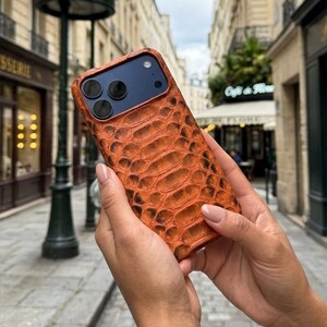 Pode incluir: Uma capa de smartphone com um padr&atilde;o texturizado de pele de cobra em tons de laranja e castanho. A capa tem um recorte para um sistema de c&acirc;mara de tr&ecirc;s lentes e um flash. A superf&iacute;cie apresenta um design de escamas em relevo no verso.