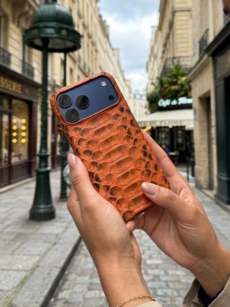 Pode incluir: Uma capa de smartphone com um padr&atilde;o texturizado de pele de cobra em tons de laranja e castanho. A capa tem um recorte para um sistema de c&acirc;mara de tr&ecirc;s lentes e um flash. A superf&iacute;cie apresenta um design de escamas em relevo no verso.