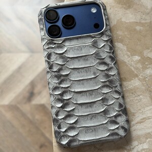 May include: A gray and white snakeskin-textured phone case. The case has a blue camera panel with three black camera lenses. The case is designed to fit a smartphone and provides protection with a stylish look.