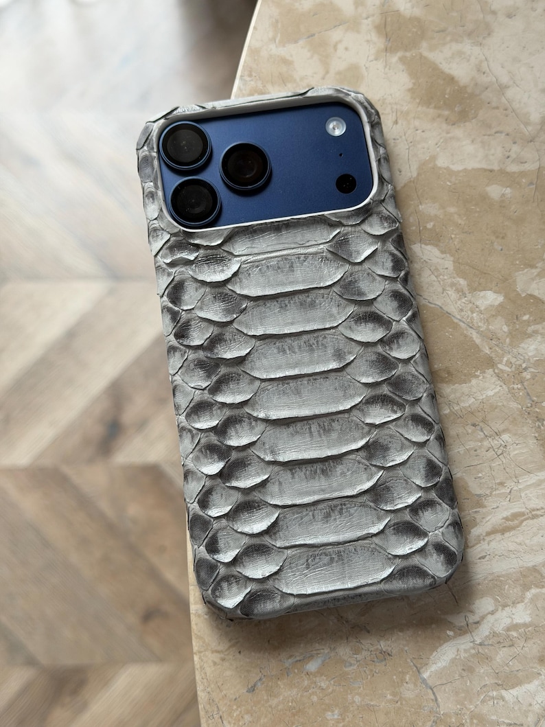 May include: A gray and white snakeskin-textured phone case. The case has a blue camera panel with three black camera lenses. The case is designed to fit a smartphone and provides protection with a stylish look.