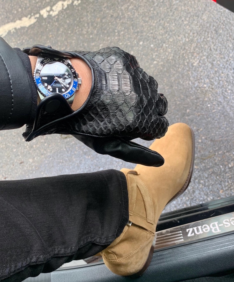May include: A black leather driving glove with a snakeskin pattern. The glove is worn on the left hand of a person wearing black jeans and tan suede boots. A silver watch with a black face and blue accents is on the wrist.