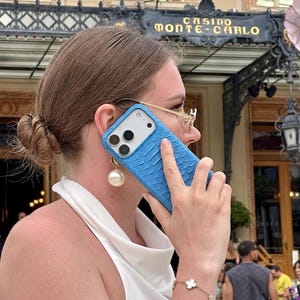 Pode incluir: Uma capa de smartphone azul brilhante com um padr&atilde;o texturizado semelhante a escamas na parte traseira. A capa apresenta um recorte para as lentes da c&acirc;mera e &eacute; mostrada encostada a uma orelha. Tem um acabamento mate e cobre o verso e lados.
