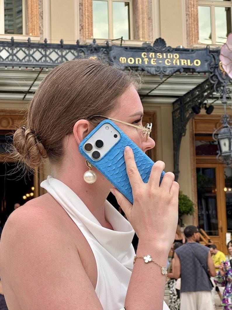 Pode incluir: Uma capa de smartphone azul brilhante com um padr&atilde;o texturizado semelhante a escamas na parte traseira. A capa apresenta um recorte para as lentes da c&acirc;mera e &eacute; mostrada encostada a uma orelha. Tem um acabamento mate e cobre o verso e lados.
