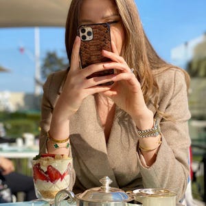 May include: A brown snakeskin-effect phone case is held up. A dessert with strawberries and cream is in a clear glass on a table with a silver teapot and teacup. The person wears a tan blazer and gold jewellery.