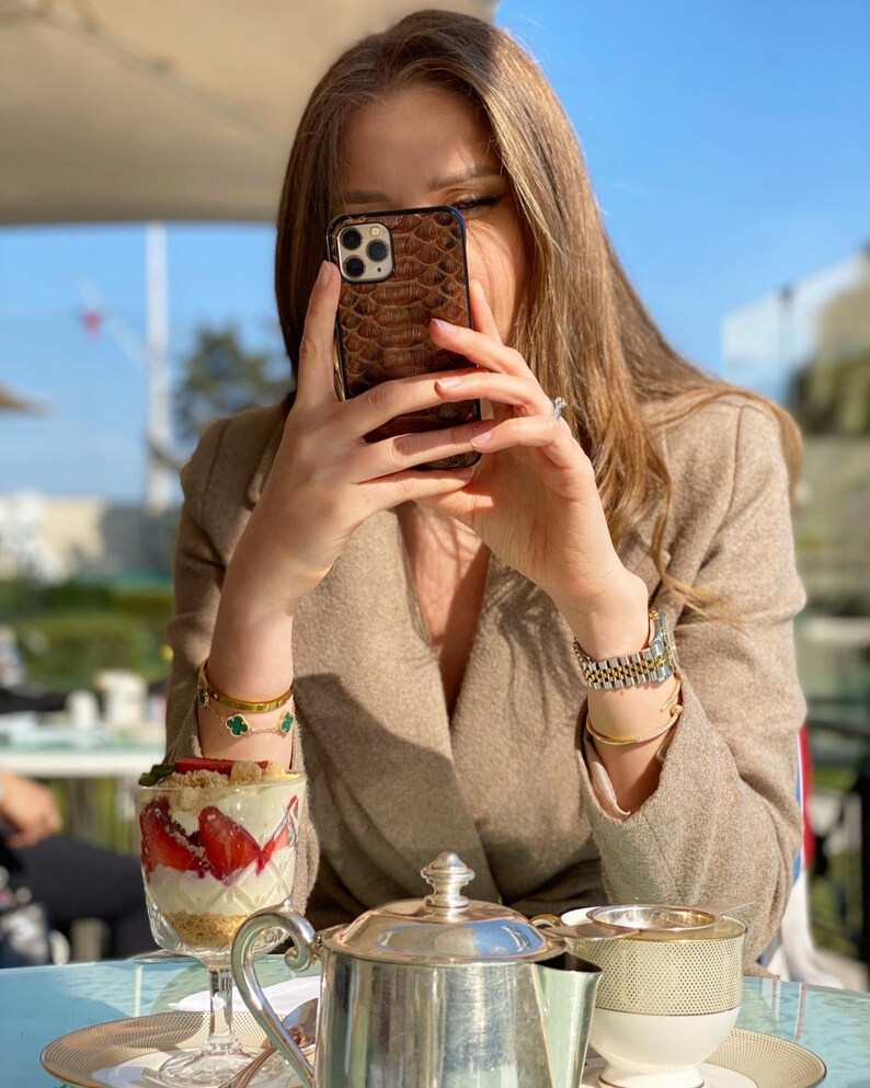 May include: A brown snakeskin-effect phone case is held up. A dessert with strawberries and cream is in a clear glass on a table with a silver teapot and teacup. The person wears a tan blazer and gold jewellery.
