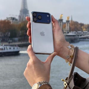Puede incluir: Un tel&eacute;fono inteligente plateado y azul con una c&aacute;mara de triple lente se sostiene frente a la Torre Eiffel. El tel&eacute;fono tiene una parte trasera plateada con un logotipo de Apple y un m&oacute;dulo de c&aacute;mara azul. El tel&eacute;fono est&aacute; en una funda plateada.
