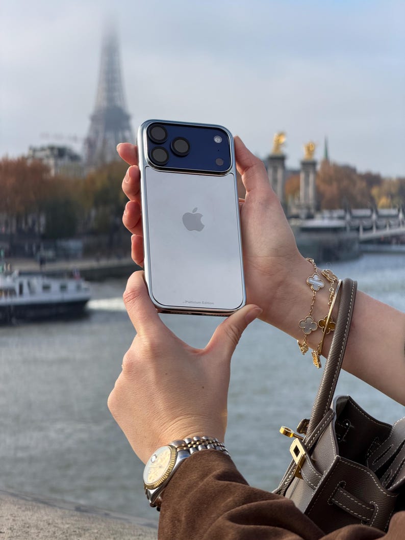 Puede incluir: Un tel&eacute;fono inteligente plateado y azul con una c&aacute;mara de triple lente se sostiene frente a la Torre Eiffel. El tel&eacute;fono tiene una parte trasera plateada con un logotipo de Apple y un m&oacute;dulo de c&aacute;mara azul. El tel&eacute;fono est&aacute; en una funda plateada.