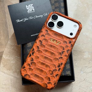 May include: An orange snakeskin-effect phone case with gold lettering spelling "SAZ". The case is presented in a black box with a card reading "Thank You For Choosing Us!" and the brand logo. The case is designed for a smartphone.
