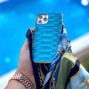 May include: A turquoise blue phone case with a scale-like pattern and a gold-coloured frame. The phone case is held in front of a blue and green patterned scarf. A rose gold watch is visible in the foreground.