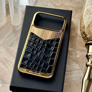 May include: A rectangular phone case featuring a polished gold-tone metal frame, a black textured crocodile-patterned inlay, and a V-shaped gold-tone section with vertical lines. The text 24k Gold Croco Edition is printed at the bottom.