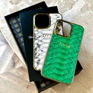 May include: Two luxury phone cases with gold trim. One case is silver with a scale pattern, the other is green with a scale pattern. Both cases have the initials "DVM" and are displayed with a black box.
