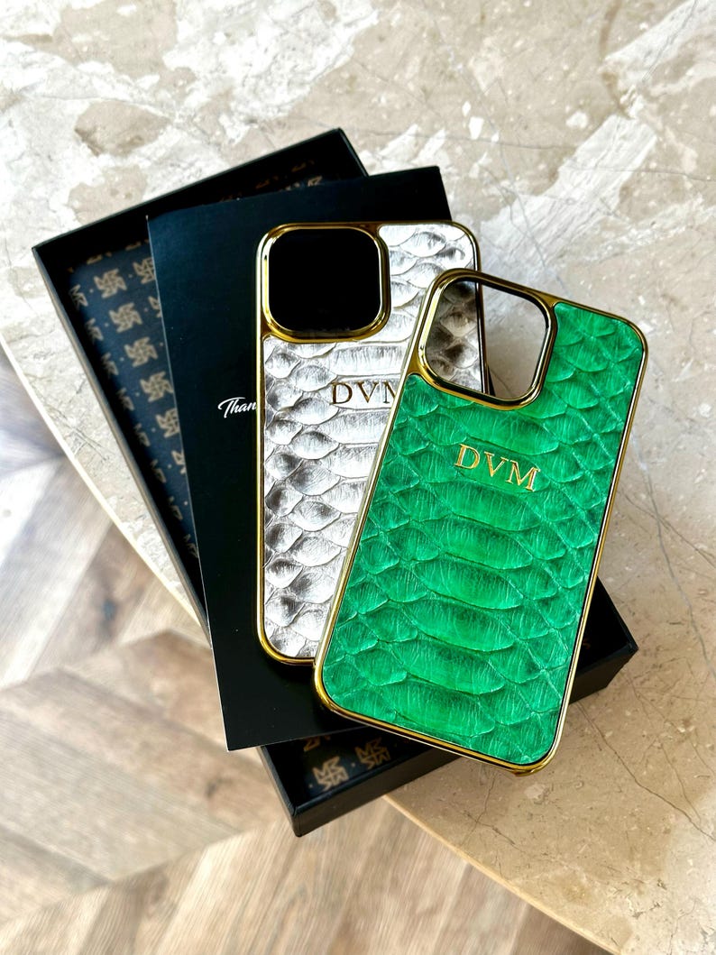 May include: Two luxury phone cases with gold trim. One case is silver with a scale pattern, the other is green with a scale pattern. Both cases have the initials "DVM" and are displayed with a black box.
