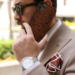 May include: A brown, textured phone case with a reptile skin pattern. The person is wearing a tan suit, sunglasses, and a wristwatch. A pocket square with a white border and red polka dots is visible. The phone is held up to the ear.