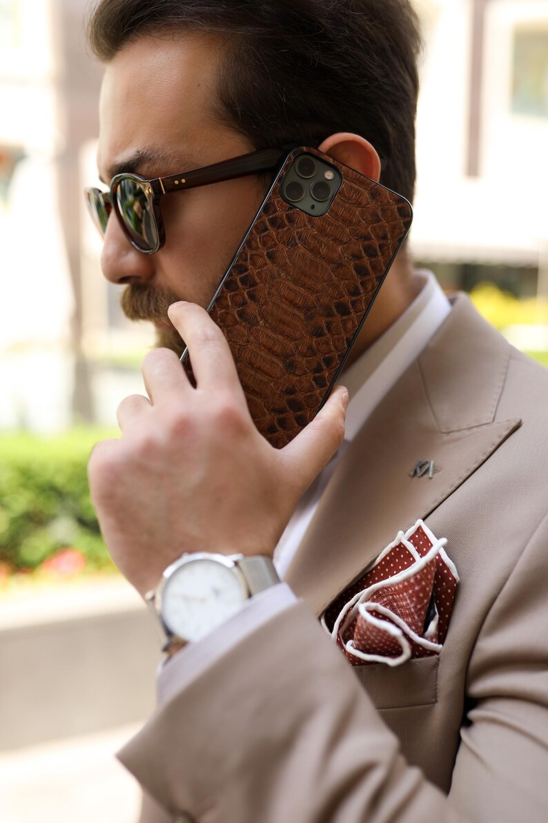 May include: A brown, textured phone case with a reptile skin pattern. The person is wearing a tan suit, sunglasses, and a wristwatch. A pocket square with a white border and red polka dots is visible. The phone is held up to the ear.
