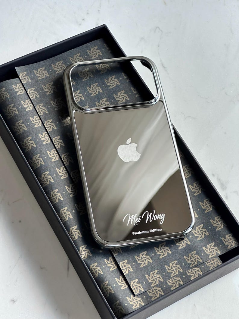 Puede incluir: Una funda de tel&eacute;fono plateada reflectante con el logotipo de Apple y el nombre "Mei Wong" y "Platinium Edition" impresos. La funda est&aacute; en una caja negra con papel estampado.