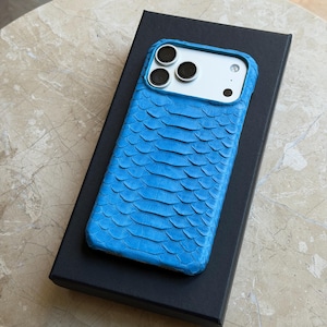 May include: A bright blue smartphone case with a textured, scale-like pattern covering the back. The case features a cutout for the camera module, revealing three circular lenses and a flash on the white back of the smartphone device.