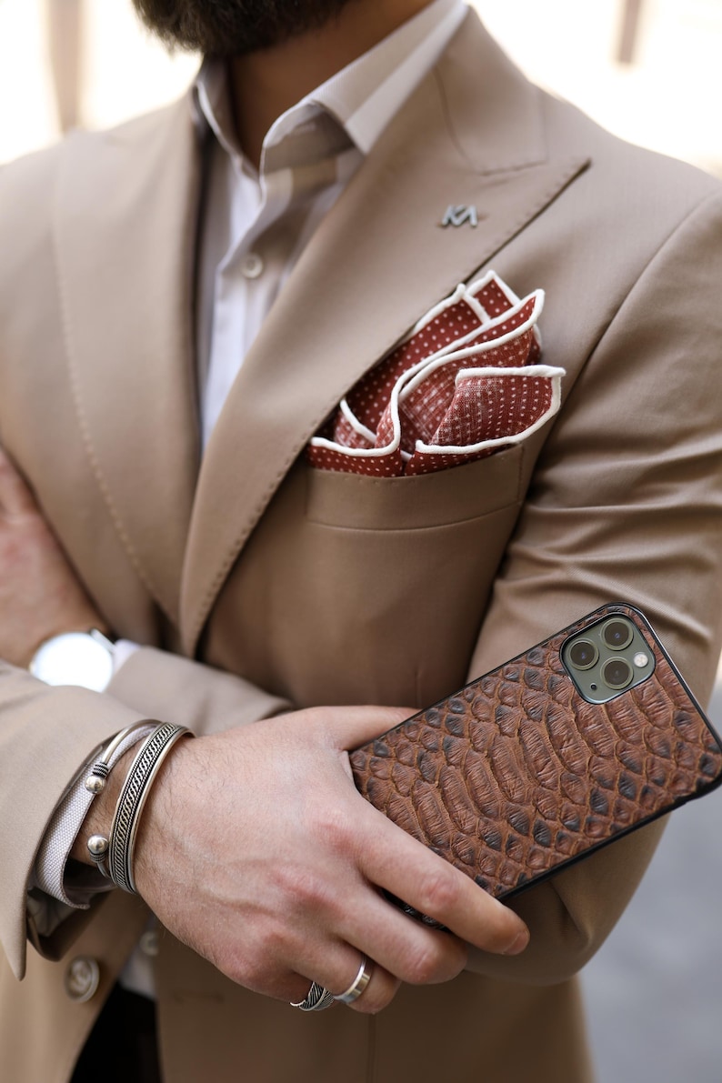 May include: A tan suit with a white shirt and a red polka dot pocket square. A brown textured phone case is held. Silver bracelets and a watch are also visible.