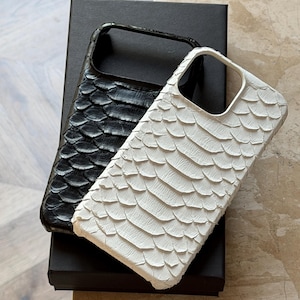 May include: Two phone cases with a textured, scaly pattern. One case is black, the other is white. The cases are resting on a black box. The cases have a cutout for the camera and buttons.