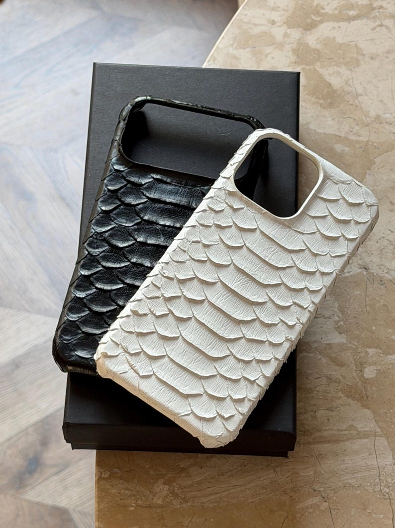 May include: Two phone cases with a textured, scaly pattern. One case is black, the other is white. The cases are resting on a black box. The cases have a cutout for the camera and buttons.