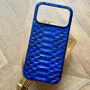 May include: A vibrant blue phone case with a textured snakeskin pattern. The case has a rectangular shape with a camera cutout and a raised edge for screen protection. The case is made of a durable material and is designed to fit a smartphone.