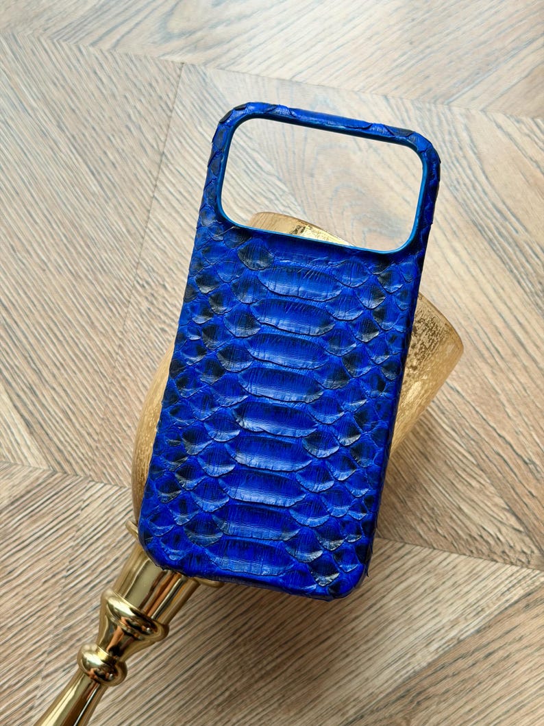 May include: A vibrant blue phone case with a textured snakeskin pattern. The case has a rectangular shape with a camera cutout and a raised edge for screen protection. The case is made of a durable material and is designed to fit a smartphone.