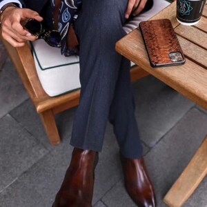 May include: A person seated outdoors wearing a navy suit, brown leather Chelsea boots, and a patterned scarf. A brown snakeskin phone case rests on a wooden table next to a cup of coffee.