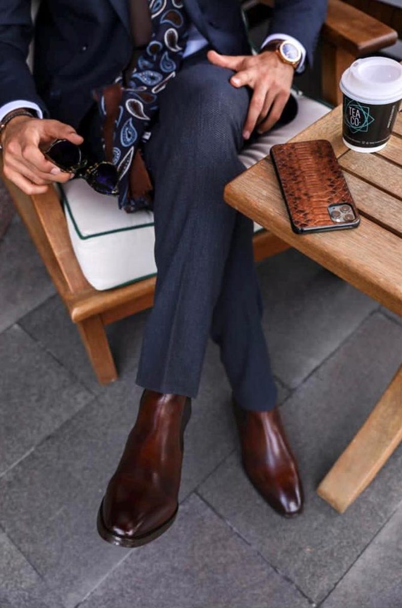 May include: A person seated outdoors wearing a navy suit, brown leather Chelsea boots, and a patterned scarf. A brown snakeskin phone case rests on a wooden table next to a cup of coffee.