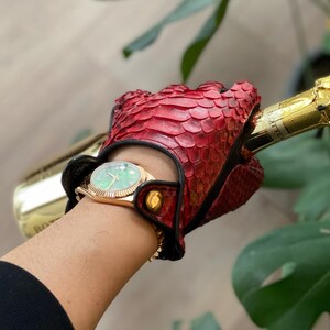 May include: A red leather driving glove with a black trim and a gold button. The glove is made of snakeskin and has a textured surface. The glove is being worn by a person holding a bottle of champagne.