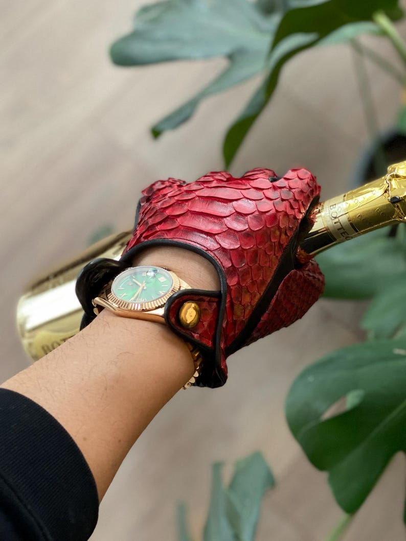 May include: A red leather driving glove with a black trim and a gold button. The glove is made of snakeskin and has a textured surface. The glove is being worn by a person holding a bottle of champagne.