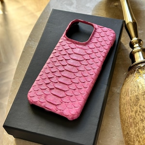 May include: A vibrant pink phone case with a textured, scale-like pattern. The case is resting on a black box, suggesting a presentation or storage context. The design is eye-catching and likely offers both style and protection.