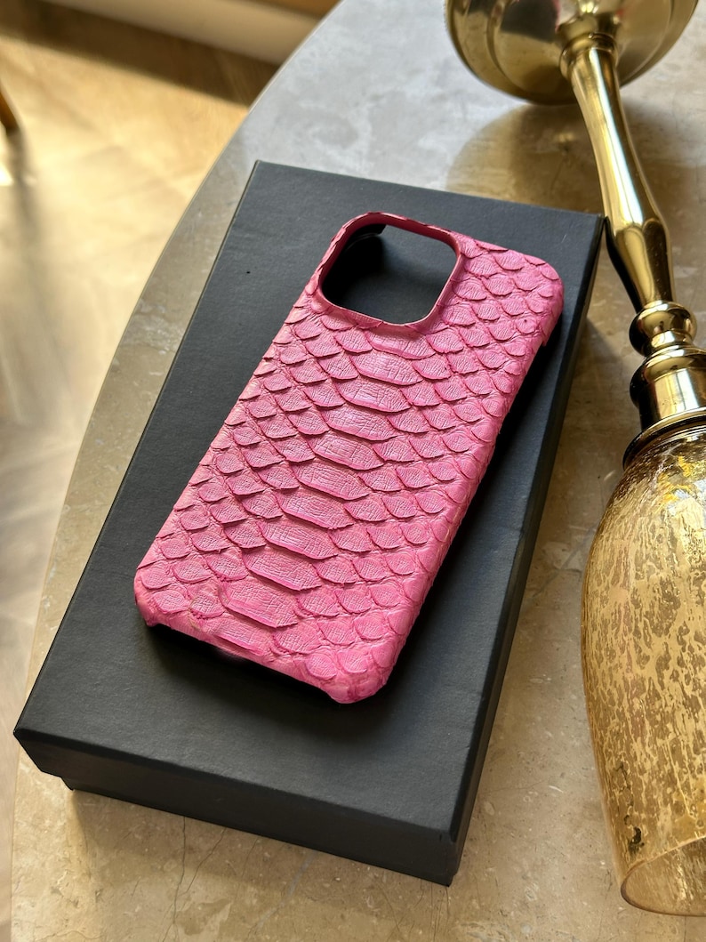 May include: A vibrant pink phone case with a textured, scale-like pattern. The case is resting on a black box, suggesting a presentation or storage context. The design is eye-catching and likely offers both style and protection.