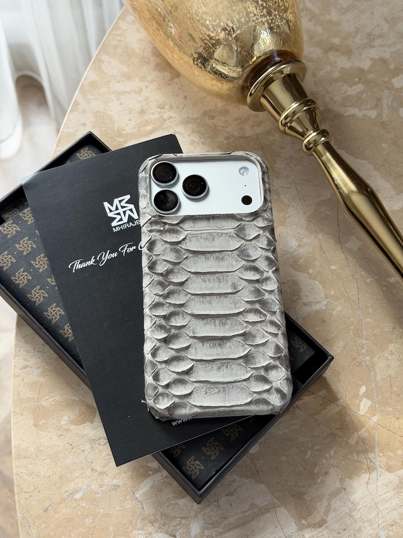 May include: A gray snakeskin-textured phone case with a triple-lens camera cutout. The case is displayed on a marble surface next to a black box containing a card with the text "Thank You For" and a logo. A gold decorative object is in the background.