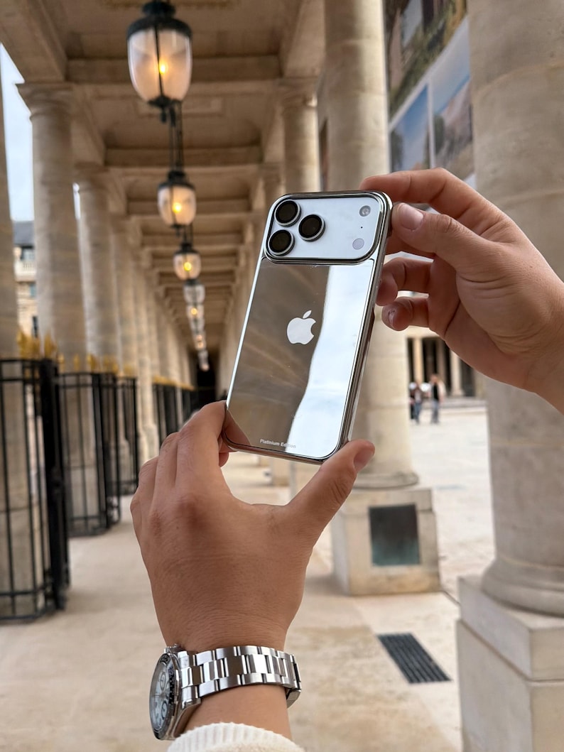 Puede incluir: Funda plateada para iPhone con superficie reflectante, con el logotipo de Apple. La funda tiene un recorte para la c&aacute;mara con tres lentes. La funda se sostiene frente a un fondo borroso de un edificio con columnas y luces.