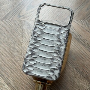 May include: A gray and white snakeskin-patterned phone case. The case has a rectangular shape with a camera cutout and a raised edge for screen protection. The case is resting on a gold-colored stand.