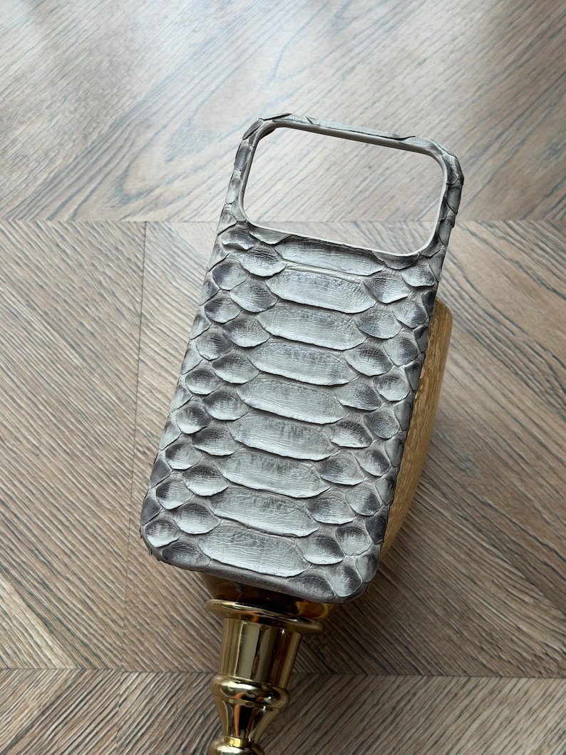 May include: A gray and white snakeskin-patterned phone case. The case has a rectangular shape with a camera cutout and a raised edge for screen protection. The case is resting on a gold-colored stand.