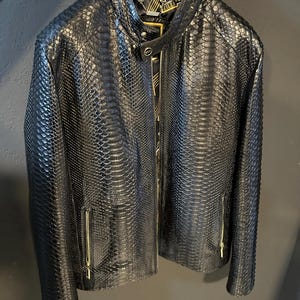 Könnte beinhalten: Schwarze Lederjacke mit Schlangenhautmuster. Die Jacke hat einen Reißverschluss und goldene Akzente.