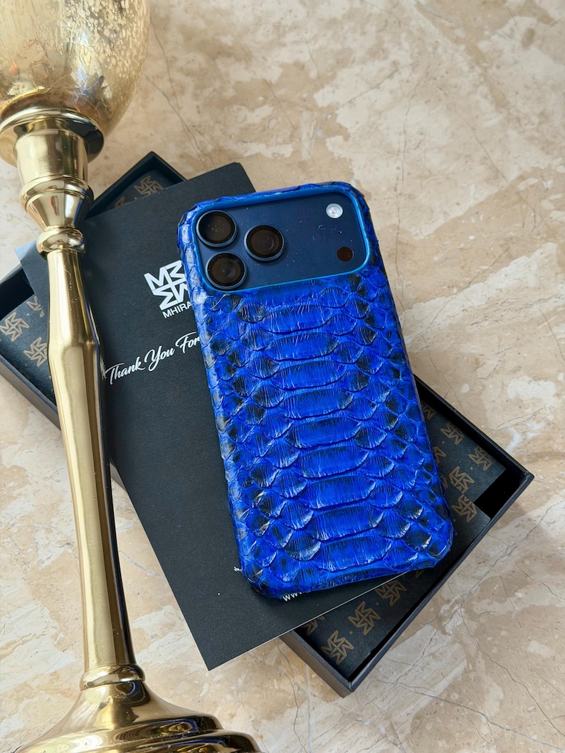 May include: A blue phone case with a snakeskin pattern. The case has a dark blue frame and three camera cutouts. The case is in a black box with the text "Thank You For" and a logo. A gold candlestick is in the background.