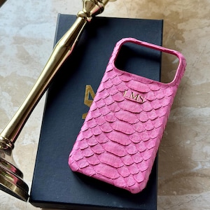 May include: A pink phone case with a snakeskin texture and the initials "LMS" in gold. The case is resting on a black box, with a gold candlestick in the background. The case is designed to protect a smartphone.