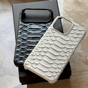 May include: Two phone cases, one black and one white, with a textured, scale-like pattern. The cases are rectangular with a cutout for the camera and are resting on a black box.