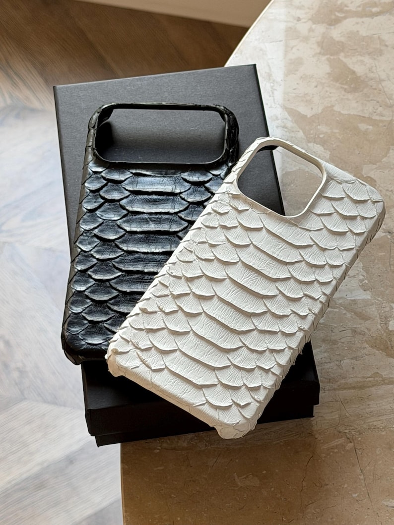 May include: Two phone cases, one black and one white, with a textured, scale-like pattern. The cases are rectangular with a cutout for the camera and are resting on a black box.
