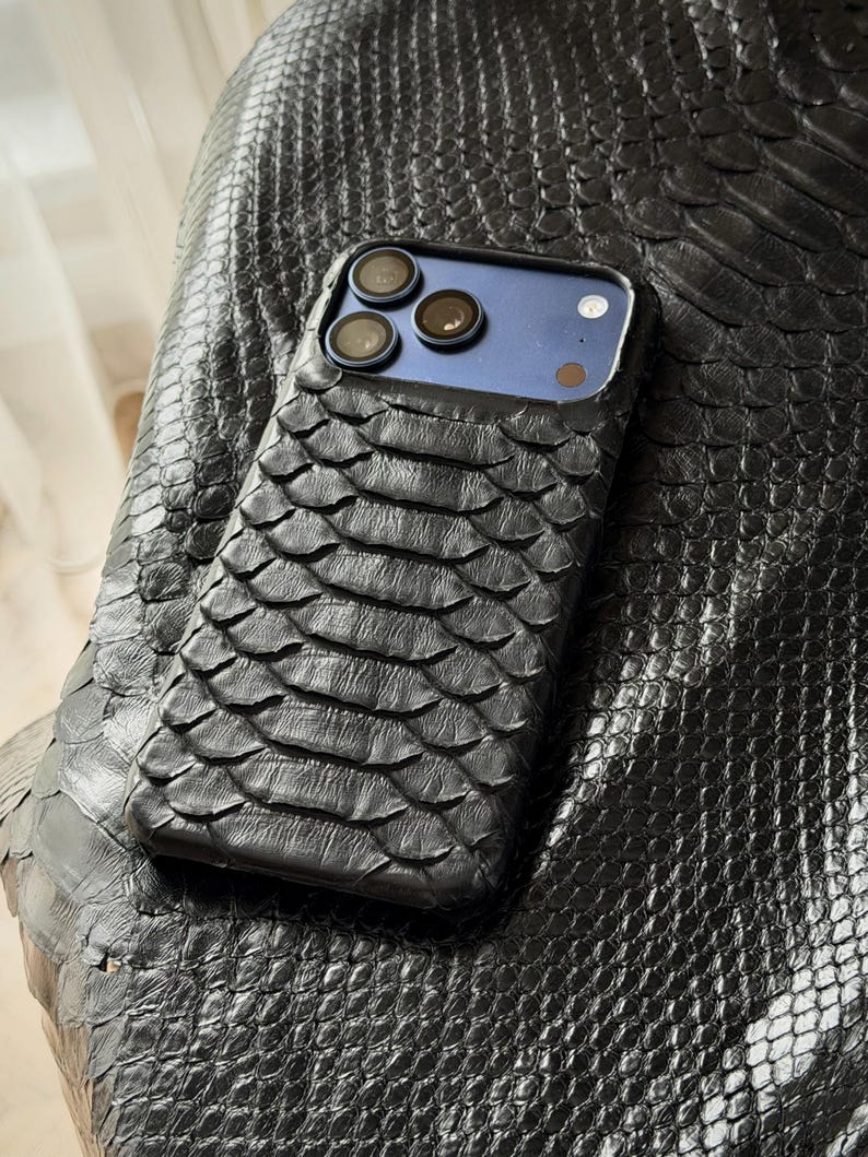 May include: A black phone case with a textured, scaly pattern, possibly leather or a similar material, rests on a matching surface. The case has a blue back and three camera lenses. The overall aesthetic is sleek and luxurious.