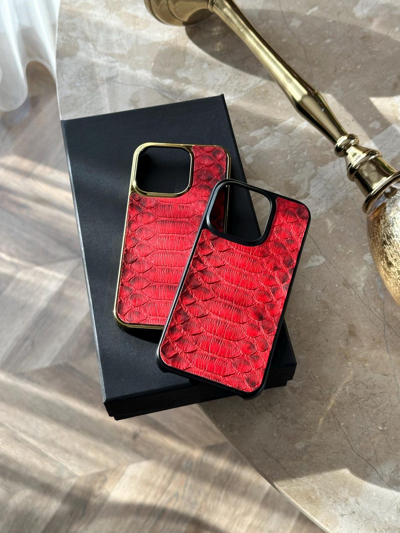 May include: Two red phone cases with a reptile skin texture. One case has a gold frame, the other has a black frame. Both cases are resting on a black box.