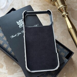 May include: A black phone case with a white border, resting in an open black box with a patterned interior. The case has a black suede-like interior. A card with the text "Thank You For" is visible.